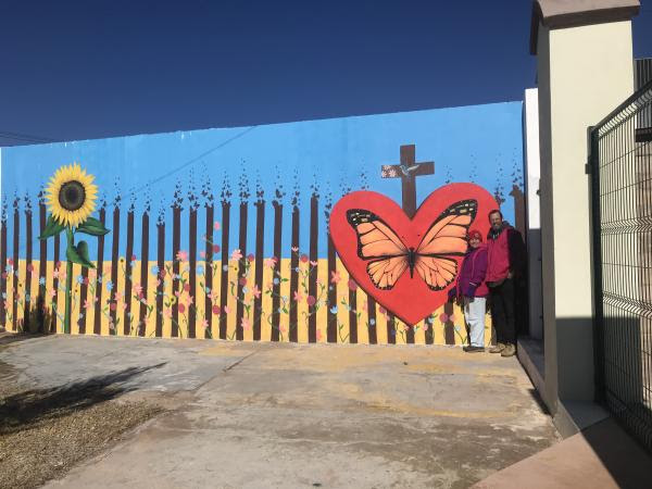 Two people stand in front of the US-Mexico border fence in Agua Prieta, painted as a mural with a butterfly in the center of a heart.